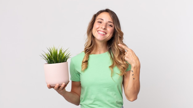 Mujer muy delgada sonriendo con confianza apuntando a su propia sonrisa amplia y sosteniendo una planta decorativa