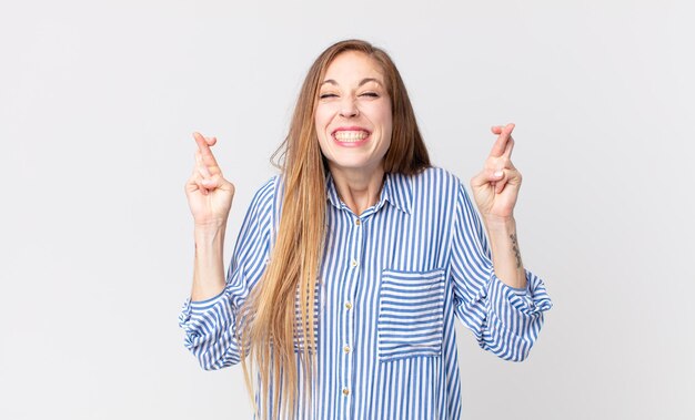 Mujer muy delgada sonriendo y cruzando ansiosamente ambos dedos, sintiéndose preocupada y deseando o esperando buena suerte