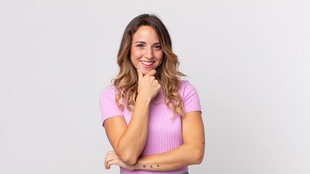 Mujer muy delgada sonriendo, disfrutando de la vida, sintiéndose feliz, amigable, satisfecha y despreocupada con la mano en la barbilla