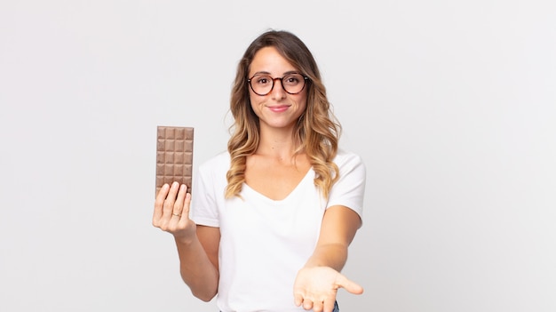 Mujer muy delgada sonriendo felizmente con amigable y ofreciendo y mostrando un concepto y sosteniendo una barra de chocolate