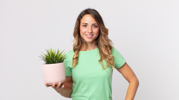 Mujer muy delgada sonriendo felizmente con una mano en la cadera y confiada y sosteniendo una planta decorativa