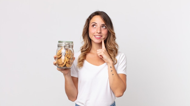 Mujer muy delgada sonriendo felizmente y soñando despierto o dudando y sosteniendo una botella de vidrio de galletas