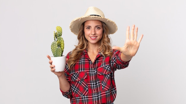 Mujer muy delgada sonriendo y mirando amigable, mostrando el número cinco sosteniendo un cactus. concepto de granjero