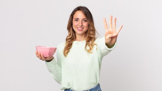 Mujer muy delgada sonriendo y mirando amigable, mostrando el número cuatro y sosteniendo una olla vacía.