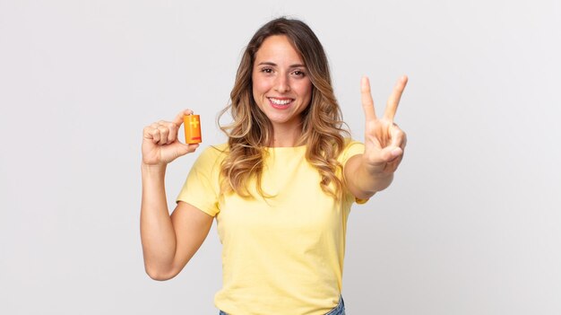 Mujer muy delgada sonriendo y mirando amigable, mostrando el número dos y sosteniendo las baterías