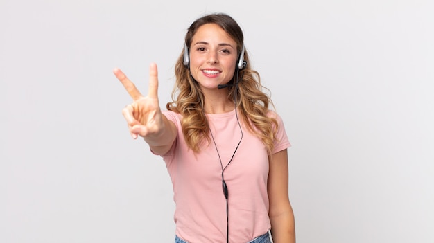 Mujer muy delgada sonriendo y mirando feliz, gesticulando victoria o paz asistente de operador con un auricular