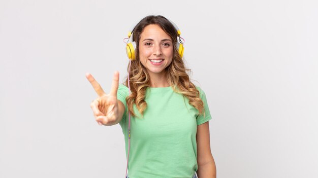 Mujer muy delgada sonriendo y mirando feliz, gesticulando victoria o paz escuchando música con auriculares