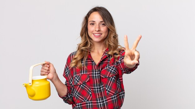 Mujer muy delgada sonriendo y mirando feliz, gesticulando victoria o paz y sosteniendo una tetera