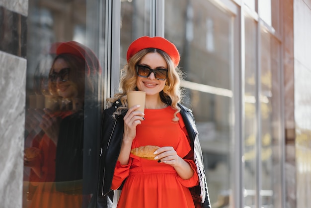 Mujer muy feliz con dulce sonrisa en gafas de sol vintage con sombrero en ropa casual rojo-negro con paseos de café y sonrisas al aire libre en la ciudad