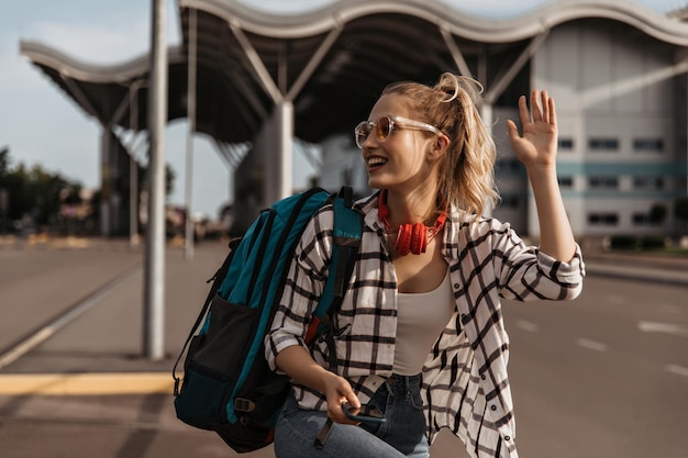 Una mujer muy guay con camisa a cuadros, top blanco y jeans, sonríe y saluda con la mano. El turista con mochila usa auriculares y posa cerca del aeropuerto.