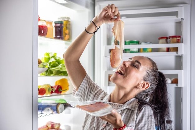 Mujer muy hambrienta en pijama disfrutando de prosciutto por la noche junto a la nevera