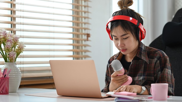 Mujer muy joven que usa auriculares inalámbricos con micrófono y podcast de audio de transmisión portátil en el estudio en casa