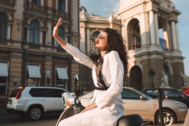 Mujer muy sonriente con cabello oscuro y rizado en traje blanco sentada en un ciclomotor y felizmente cubriendo su rostro con la mano de la luz del sol con vista a la ciudad en el fondo