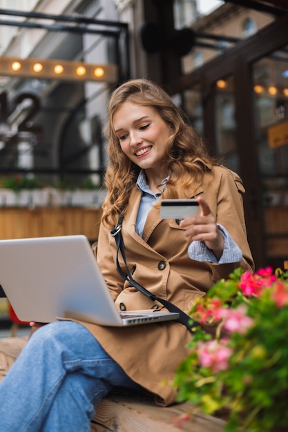 Mujer muy sonriente en gabardina trabajando con alegría en la computadora portátil mientras sostiene la tarjeta de crédito en la mano al aire libre en la terraza del café