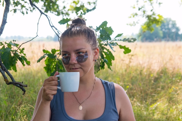 Una mujer en la naturaleza cuida su rostro. Bebe té café en la naturaleza de vacaciones.