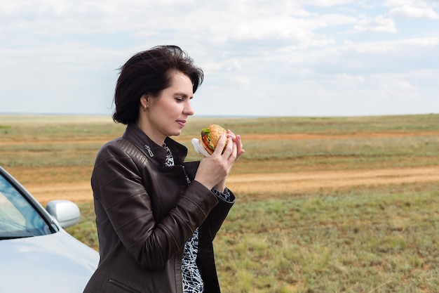 Una mujer en la naturaleza con un vaso en la mano se va a comer una hamburguesa.