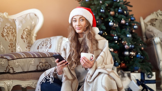 Mujer de Navidad vestida con suéter blanco sombrero de Santa en el suelo cerca del árbol de Navidad con smartpone y