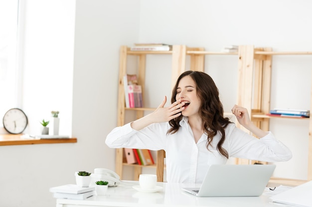 Mujer de negocios aburrida sentada en el escritorio con los ojos cerrados y bostezando Joven mujer de negocios caucásica en su oficina
