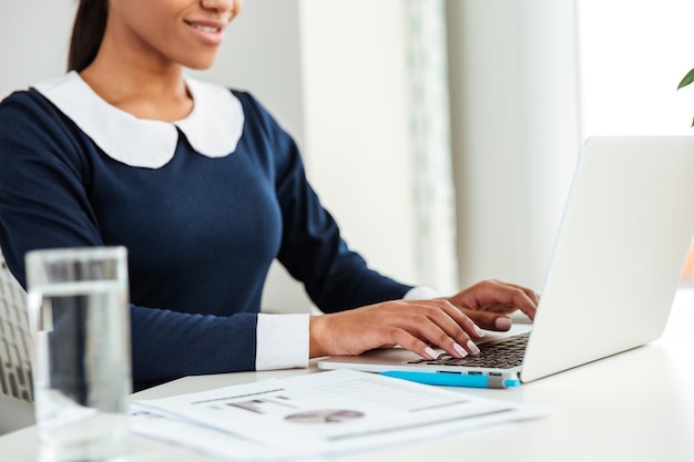 Foto mujer de negocios africana joven en vestido sentado junto a la mesa en el lugar de trabajo con agua y usando la computadora portátil. imagen recortada