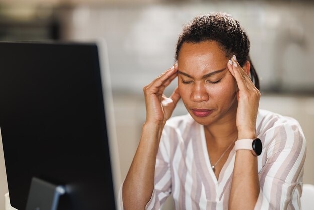 Mujer de negocios africana que sufre dolor de cabeza y experimenta estrés en el trabajo mientras trabaja en su computadora en casa.