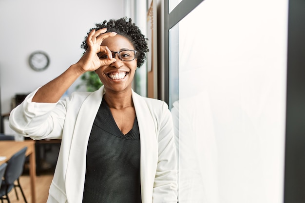 Mujer de negocios africana que trabaja en la oficina sonriendo feliz haciendo el signo de ok con la mano mirando a través de los dedos