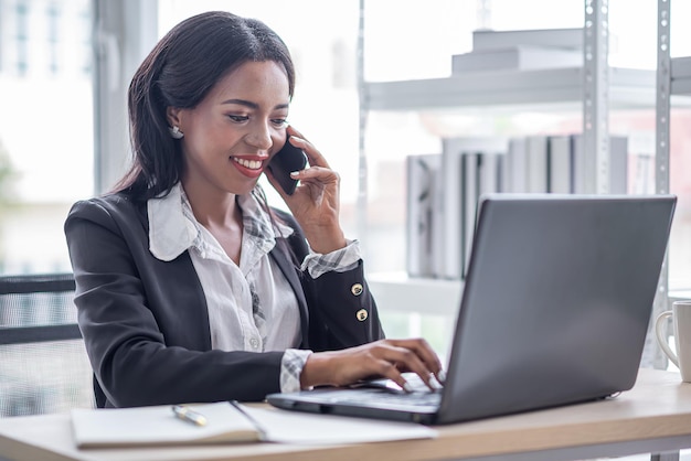 Mujer de negocios africana que trabaja en la oficina usando una conversación de computadora por teléfono inteligente con el cliente