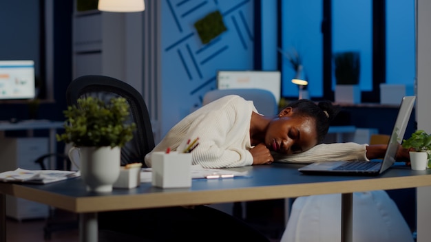 Mujer de negocios africana de sobrecarga agotada que se queda dormida en el escritorio con el monitor de la computadora portátil abierta mientras trabaja en la puesta en marcha de la empresa. Empleado con exceso de trabajo que utiliza haciendo horas extraordinarias respetando el plazo, durmiendo
