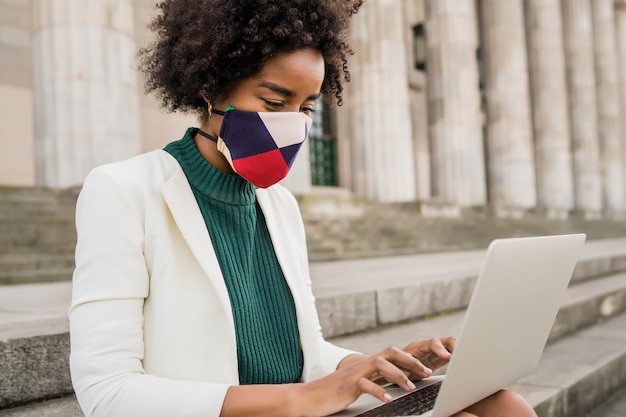 Mujer de negocios afro con máscara protectora y usando su computadora portátil mientras está sentada en las escaleras al aire libre