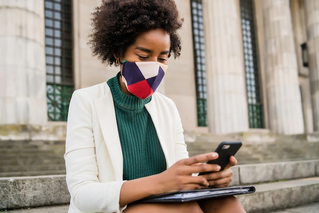 Mujer de negocios afro con máscara protectora y usando su teléfono móvil mientras está sentado en las escaleras al aire libre en la calle