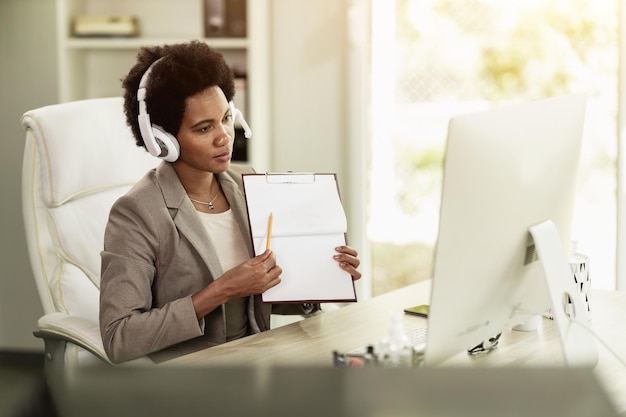 Mujer de negocios afroamericana con auriculares con videollamada mientras está sentada sola en una oficina en el trabajo.