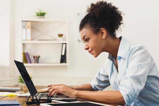 Mujer de negocios afroamericana feliz trabajando en equipo portátil en la oficina. La empresaria escribiendo algo en la computadora mientras está sentada en su lugar de trabajo, vista lateral, espacio de copia