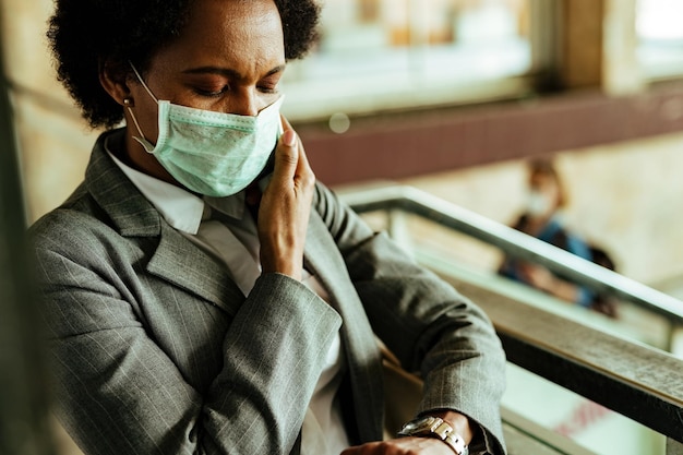 Mujer de negocios afroamericana con máscara protectora y control de tiempo en reloj de pulsera mientras está en la estación de tren pública