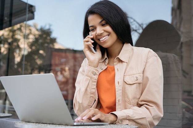 Mujer de negocios afroamericana trabajando en línea, utilizando equipo portátil, hablando por teléfono móvil sentado en la cafetería