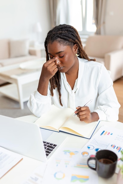 Mujer de negocios agotada que tiene dolor de cabeza en la oficina en casa Mujer creativa afroamericana que trabaja en el escritorio de la oficina sintiéndose cansada Mujer de negocios estresada que siente dolor en los ojos mientras trabaja demasiado