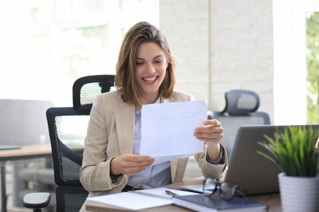 Mujer de negocios alegre atractiva que controla los documentos en papel en la oficina, trabajando en la computadora portátil.