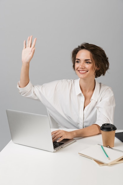 Mujer de negocios alegre feliz posando aislado sobre pared gris sentado en la mesa usando laptop