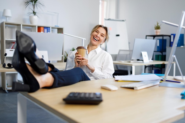 Foto mujer de negocios alegre con una taza de café poniendo piernas sobre la mesa en la oficina