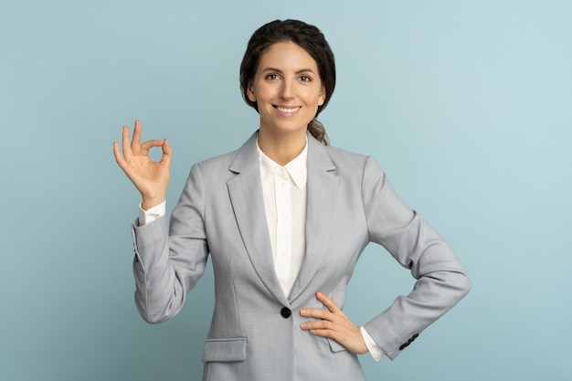 Mujer de negocios alegre usa chaqueta de buen humor sonriendo ampliamente, mostrando signo de ok sobre fondo de color azul de estudio. Empleada de oficina positiva anuncia un producto. Cliente, venta, descuento.