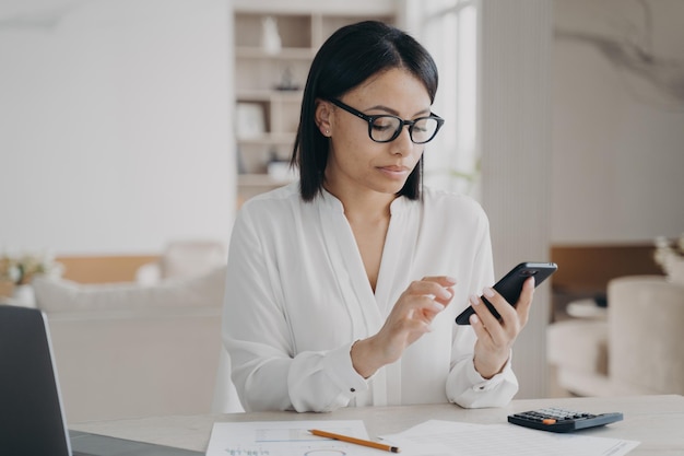 Mujer de negocios con anteojos usando aplicaciones de teléfonos móviles para la gestión del tiempo de negocios sentada en una laptop