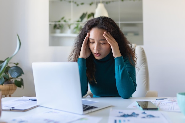 Mujer de negocios asiática agotada que tiene dolor de cabeza en la oficina Mujer creativa que trabaja en el escritorio de la oficina sintiéndose cansada Mujer de negocios informal estresada que siente dolor de cabeza mientras trabaja demasiado en la computadora portátil