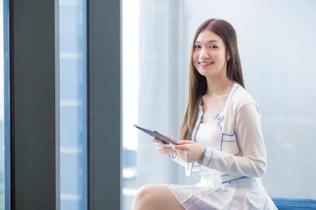 Mujer de negocios asiática con camisa blanca sostiene una tableta mientras se sienta en una silla en la oficina del lugar de trabajo.