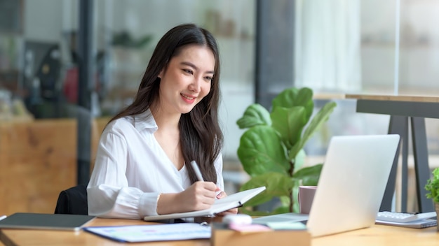 Mujer de negocios asiática confiada sentada y tomando notas con una computadora portátil en la oficina felizmente.