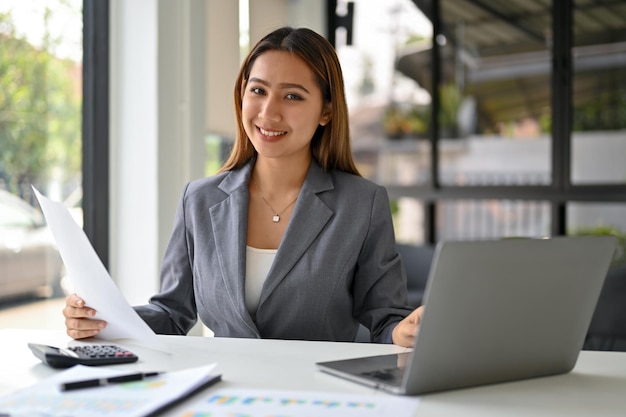 Una mujer de negocios asiática confiada se sienta en su escritorio con una computadora portátil e informes financieros