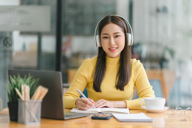 Una mujer de negocios asiática enfocada con auriculares está tomando notas en un cuaderno mientras ve un curso de video webinar Una estudiante seria escuchando la conferencia para estudiar en línea a través del aprendizaje electrónico