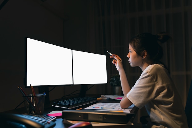 Mujer de negocios asiática estresante sentado en la silla y mirar las dos computadoras con pantalla en blanco en la oficina por la noche.