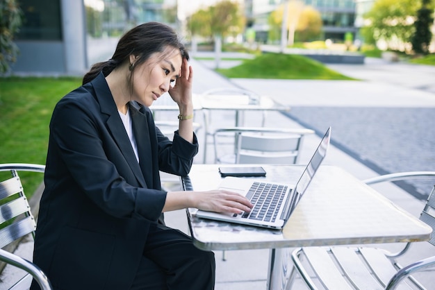 Mujer de negocios asiática con exceso de trabajo cansada en traje con computadora portátil trabajando al aire libre