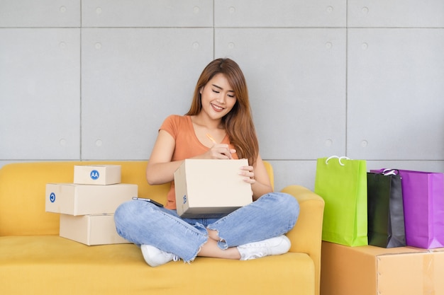 La mujer de negocios asiática feliz hermosa joven con la cara sonriente está escribiendo el nombre y la dirección del cliente en un empaquetado de la caja del paquete