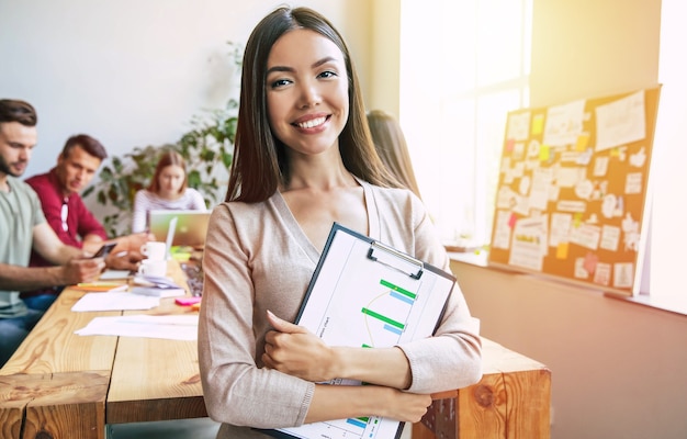 La mujer de negocios asiática hermosa confiada joven feliz en ropa de sport elegante está planteando en el fondo del equipo de negocios moderno. Compañeros de trabajo. Poner en marcha el equipo. Estudiantes juntos