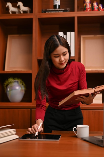Mujer de negocios asiática inteligente buscando información sobre el libro y usando una tableta digital
