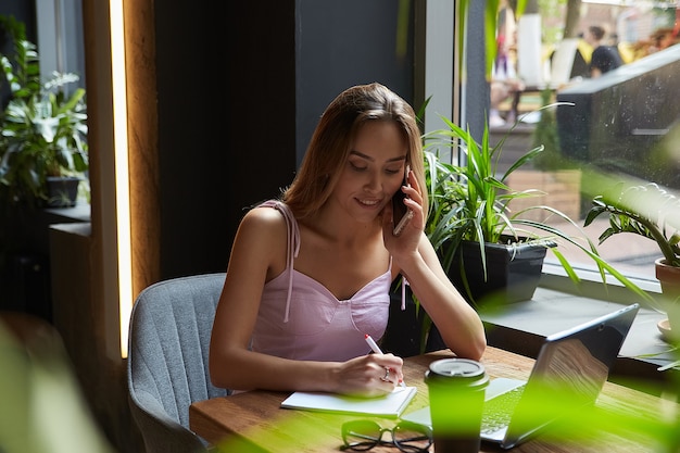 Mujer de negocios asiática joven que escribe notas en el cuaderno en el café que trabaja en la computadora portátil